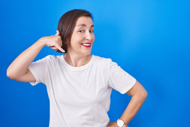 Middelbare leeftijd Latijns-Amerikaanse vrouw die over een blauwe achtergrond glimlacht doen telefoon gebaar met de hand en vingers als praten aan de telefoon. communicatie van concepten.  - Foto, afbeelding