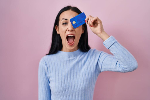 Hispanische Frau mit Kreditkarte über den Augen wütend und wütend schreiend frustriert und wütend, schreiend vor Wut. Wut und aggressives Konzept.  - Foto, Bild