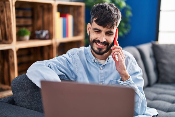 Nuori latino mies käyttää kannettavaa tietokonetta ja puhuu älypuhelimella kotona - Valokuva, kuva