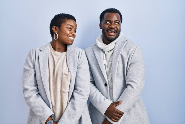 Fiatal afro-amerikai házaspár áll a kék háttér felett, együtt néztek félre mosollyal az arcukon, természetes kifejezéssel. nevetés magabiztos.  - Fotó, kép