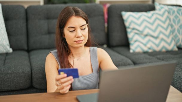 Young beautiful hispanic woman shopping with laptop and credit card sitting on floor at home - Photo, Image