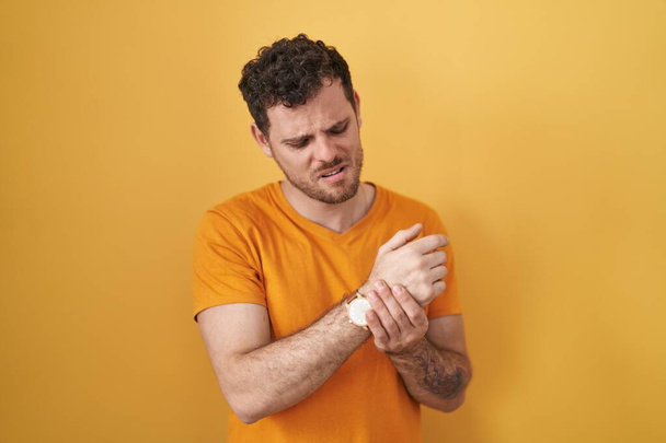 Homem hispânico jovem em pé sobre fundo amarelo sofrendo dor nas mãos e dedos, artrite inflamação  - Foto, Imagem