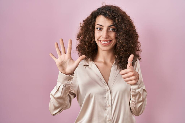 Pembe arka planda duran kıvırcık saçlı İspanyol kadın altı numaralı parmaklarını gösterip işaret ederken kendinden emin ve mutlu gülümsüyor..  - Fotoğraf, Görsel