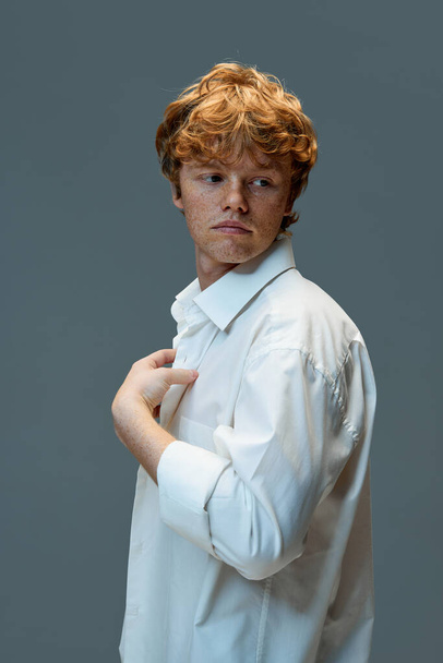 Serious young red and curly hair male in white t-shirt looking away while standing against grey background. Concept of natural beauty, fashion, self care, masculinity. Ad - Zdjęcie, obraz