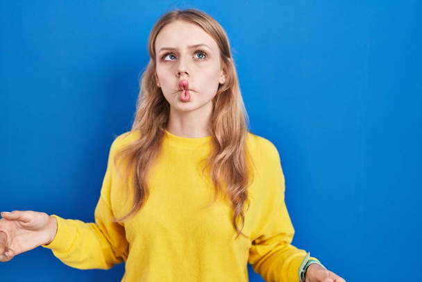 Jeune femme caucasienne debout sur fond bleu faisant visage de poisson avec des lèvres, geste fou et comique. expression drôle.  - Photo, image