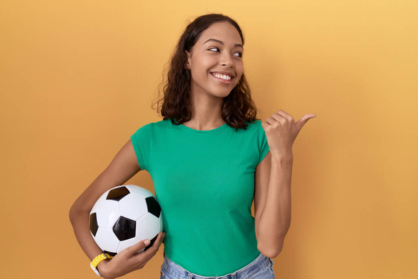 Młoda Latynoska kobieta trzymająca piłkę uśmiechnięta ze szczęśliwą twarzą patrzącą i wskazującą na bok z kciukiem w górze.  - Zdjęcie, obraz