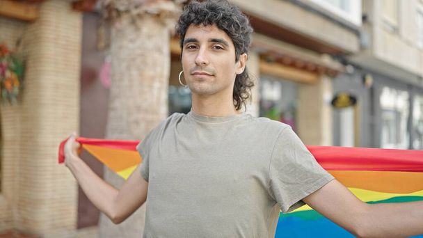 Joven hispano sosteniendo bandera de arco iris con expresión relajada en la calle - Foto, Imagen