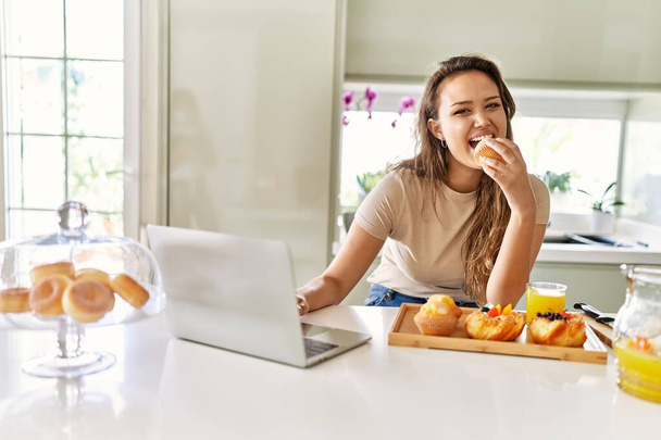 Genç ve güzel İspanyol kadın kahvaltısını mutfakta dizüstü bilgisayar kullanarak yapıyor. - Fotoğraf, Görsel
