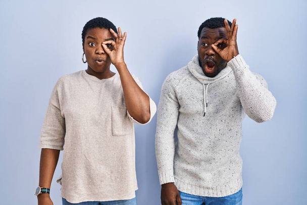 Ein junges afrikanisch-amerikanisches Paar, das vor blauem Hintergrund steht und seine Geste tut, ist schockiert mit einem überraschten Gesicht und einem Blick durch die Finger. Ungläubiger Ausdruck.  - Foto, Bild
