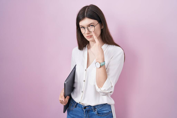Молодая белая женщина, держащая ноутбук, указывая на глаз, наблюдая за твоим жестом, подозрительным выражением  - Фото, изображение