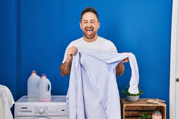 Καυκάσιος μεσήλικας που κρατάει πουκάμισο με ιδρώτα να βγάζει τη γλώσσα του χαρούμενη με αστεία έκφραση.  - Φωτογραφία, εικόνα