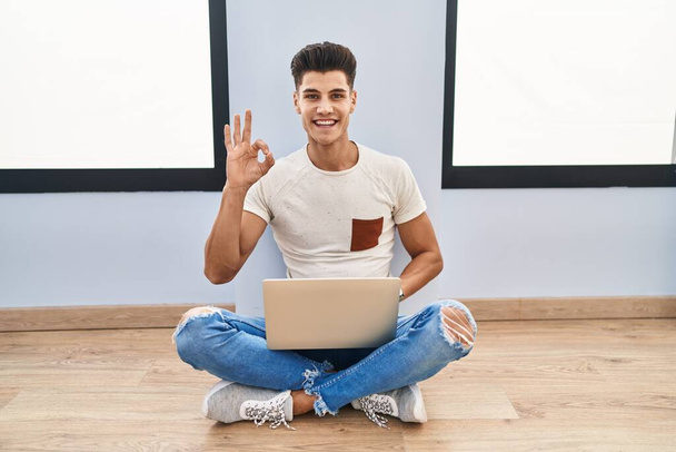 Young hispanic man using laptop at home smiling positive doing ok sign with hand and fingers. successful expression.  - Valokuva, kuva