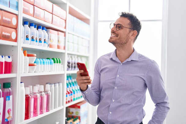 Молодий іспаномовний чоловік клієнт посміхається впевнено використовуючи смартфон в аптеці
 - Фото, зображення