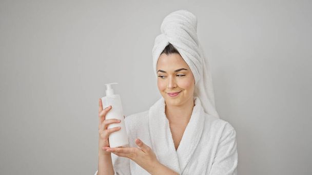 Młoda piękna Latynoska kobieta nosi szlafrok trzymając butelkę balsamu na białym tle - Zdjęcie, obraz