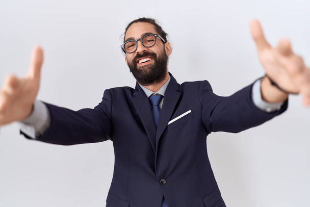 Homem hispânico com barba vestindo terno e gravata olhando para a câmera sorrindo de braços abertos para abraço. expressão alegre abraçando a felicidade.  - Foto, Imagem