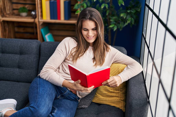 Mladá krásná hispánská žena čtení knihy sedí na pohovce doma - Fotografie, Obrázek