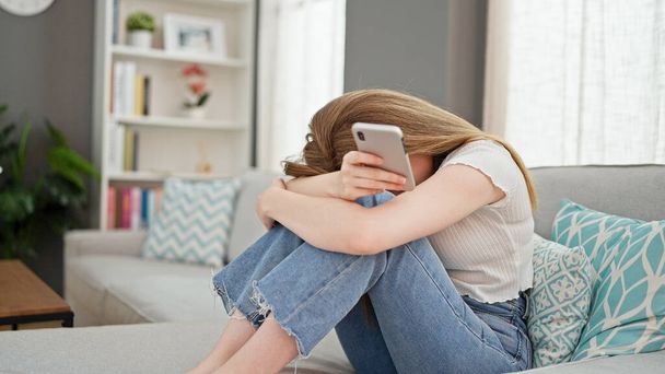 Mujer rubia joven usando teléfono inteligente sentado en el sofá estresado en casa - Foto, Imagen