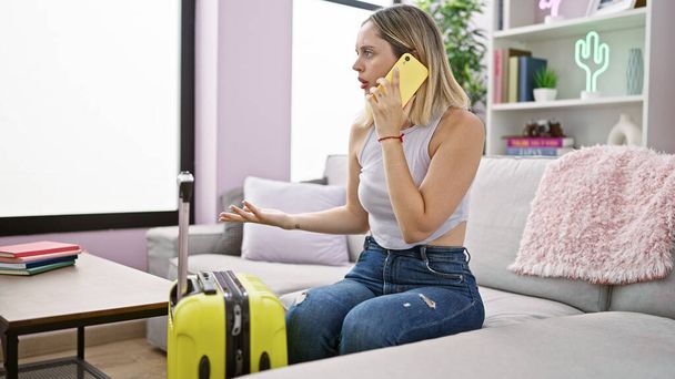 Mujer rubia joven discutiendo por teléfono inteligente sentado en el sofá por la maleta estresada en casa - Foto, imagen