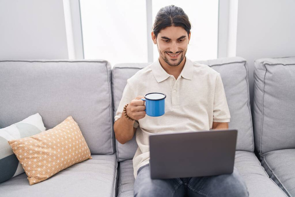 Young hispanic man using laptop and drinking coffee sitting on sofa at home - Zdjęcie, obraz