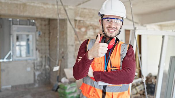Junger hispanischer Bauarbeiter lächelt selbstbewusst und macht Daumen-hoch-Geste auf Baustelle - Foto, Bild