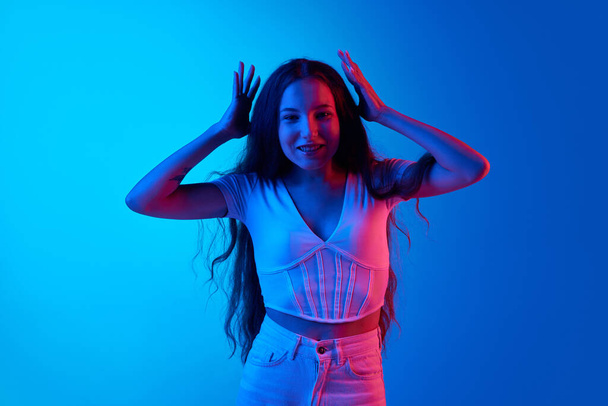 Young woman feeling cheerful and astonished, standing against blue studio background in neon light. Good and excited. Concept of human emotions, fashion, beauty, lifestyle, youth, ad - Φωτογραφία, εικόνα
