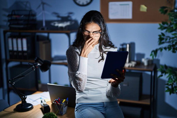 Mujer brasileña joven usando touchpad por la noche trabajando en la oficina oliendo algo apestoso y asqueroso, olor intolerable, conteniendo la respiración con los dedos en la nariz. mal olor  - Foto, Imagen