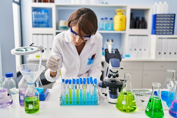 Jonge mooie Spaanse vrouw wetenschapper gieten vloeistof op reageerbuizen in het laboratorium - Foto, afbeelding