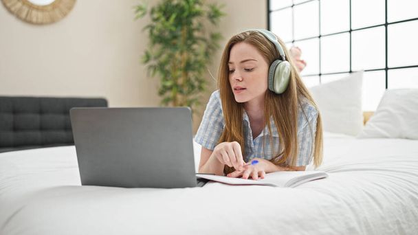 Junge blonde Studentin schreibt mit Laptop auf Notizbuch und lernt auf Bett im Schlafzimmer - Foto, Bild