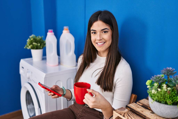 Young beautiful hispanic woman using smartphone drinking coffee waiting for washing machine at laundry room - Foto, afbeelding