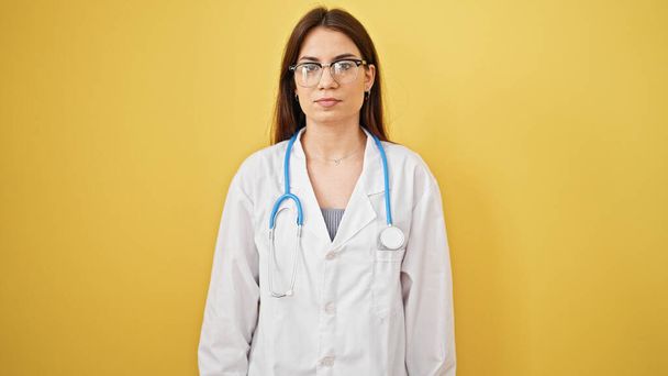 Junge schöne hispanische Ärztin steht mit ernstem Gesichtsausdruck vor isoliertem gelben Hintergrund - Foto, Bild