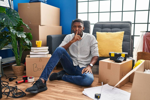 Un afroamericano seduto sul pavimento di una nuova casa che chiede di stare zitto con un dito sulle labbra. silenzio e concetto segreto.  - Foto, immagini