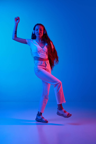 Full-length of young girl in casual clothes listening to music in headphones and dancing against blue studio background in neon light. Concept of human emotions, fashion, beauty, lifestyle, youth, ad - Photo, image