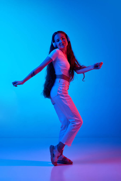Full-length of young girl in casual clothes listening to music in headphones and dancing against blue studio background in neon light. Concept of human emotions, fashion, beauty, lifestyle, youth, ad - Φωτογραφία, εικόνα