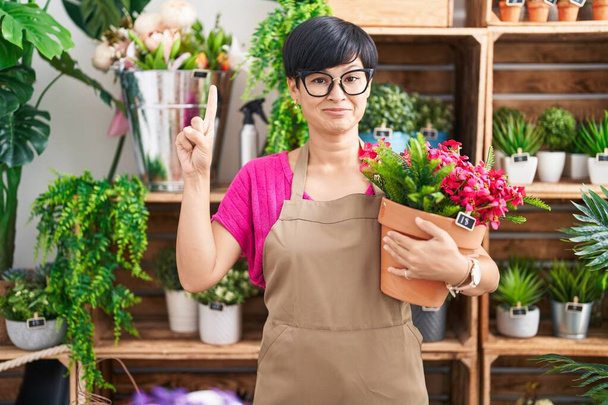 Молодая азиатка с короткими волосами, работающая в цветочном магазине, держа растение улыбающимся с идеей или вопросом, указывая пальцем со счастливым лицом, номер один  - Фото, изображение