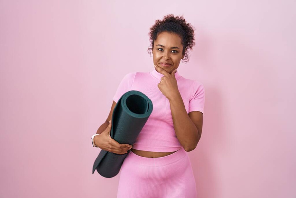 Junge hispanische Frau mit lockigem Haar, Yogamatte über rosa Hintergrund haltend und selbstbewusst in die Kamera lächelnd mit verschränkten Armen und erhobener Hand am Kinn. Positives Denken.  - Foto, Bild