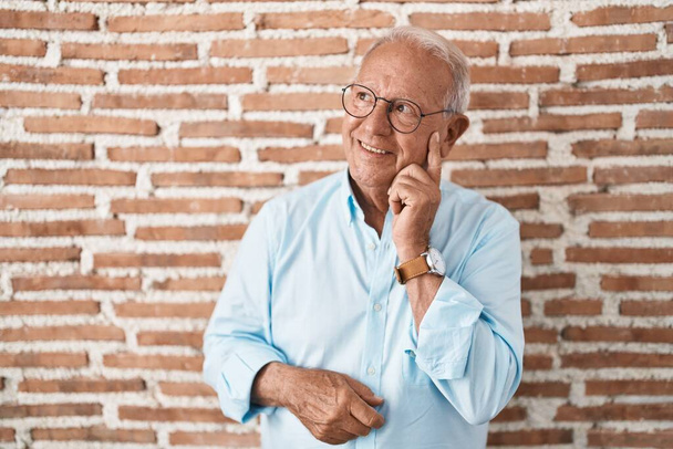 Старший мужчина с седыми волосами стоит над кирпичной стеной, думая о вопросе, обеспокоенный и нервный с рукой на подбородке  - Фото, изображение
