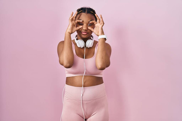 Mujer afroamericana con trenzas que usan ropa deportiva y auriculares tratando de abrir los ojos con los dedos, soñolienta y cansada de fatiga matutina  - Foto, imagen