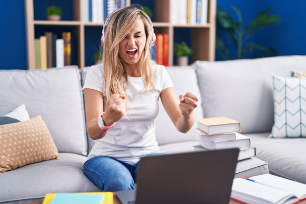 Νεαρή ξανθιά γυναίκα σπουδάζει χρησιμοποιώντας φορητό υπολογιστή στο σπίτι θυμωμένη και θυμωμένη υψώνοντας τις γροθιές και εξοργισμένη φωνάζοντας με θυμό. οργή και επιθετική αντίληψη.  - Φωτογραφία, εικόνα