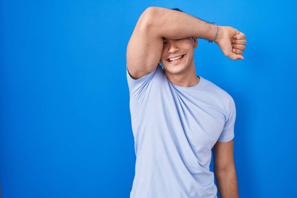 Jonge Spaanse man die over een blauwe achtergrond staat en ogen bedekt met een glimlachende arm, vrolijk en grappig. blind concept.  - Foto, afbeelding