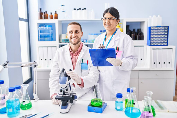 Laboratuvarda çalışan iki genç insan pozitif ve mutlu görünüyorlar. Dişlerini gösteren bir gülümsemeyle ayakta duruyorlar.  - Fotoğraf, Görsel