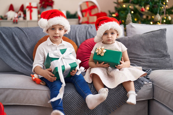 Duas crianças segurando presentes sentados no sofá pela árvore de natal em casa - Foto, Imagem