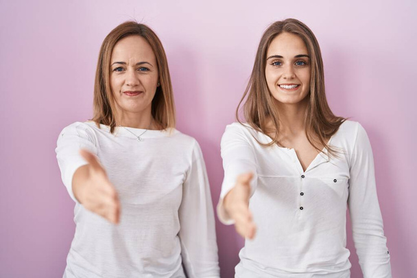 Средневековая мать и молодая дочь стоят на розовом фоне, улыбаясь дружелюбно предлагая рукопожатие, как приветствие и приветствие. успешный бизнес.  - Фото, изображение