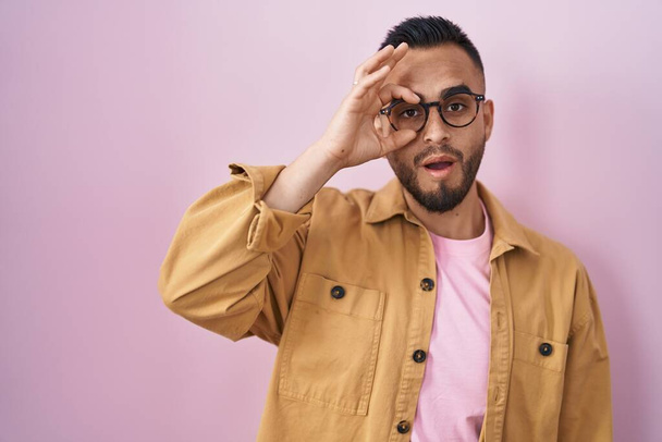 Young hispanic man standing over pink background doing ok gesture shocked with surprised face, eye looking through fingers. unbelieving expression.  - Φωτογραφία, εικόνα