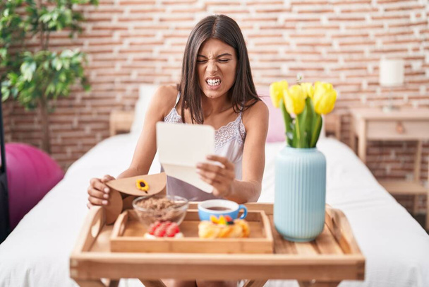 Brunette young woman eating breakfast in the bed reading a letter angry and mad screaming frustrated and furious, shouting with anger looking up.  - Photo, Image