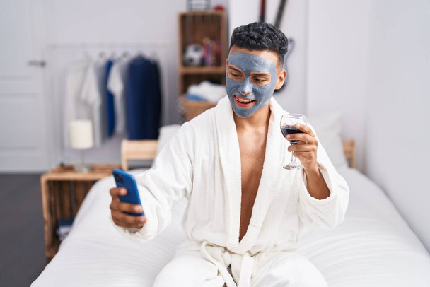 Mladý latin muž sedí na posteli, aby selfie smartphone pití vína v ložnici - Fotografie, Obrázek