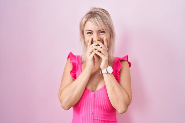 Νεαρή καυκάσια γυναίκα στέκεται πάνω από ροζ φόντο γελώντας και ντροπιασμένη χαχανίζοντας καλύπτοντας το στόμα με χέρια, κουτσομπολιά και σκανδαλώδη έννοια  - Φωτογραφία, εικόνα