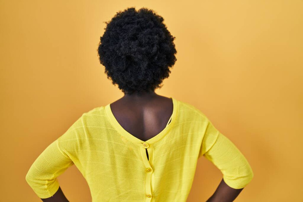 Africano jovem mulher de pé sobre estúdio amarelo de pé para trás olhando para longe com os braços no corpo  - Foto, Imagem