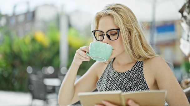 Jonge blonde vrouw drinken kopje koffie lezen boek bij coffeeshop terras - Foto, afbeelding