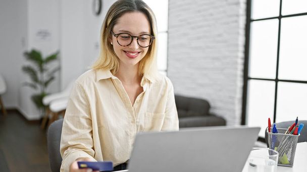 Junge blonde Geschäftsfrau kauft mit Laptop und Kreditkarte im Büro ein - Foto, Bild