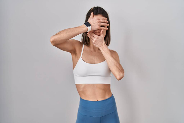 Испанская женщина в спортивной одежде на изолированном фоне, закрывая глаза и рот руками, удивлена и шокирована. скрывая эмоции  - Фото, изображение